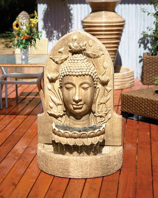 A Buddha head statue is mounted on a wall, with water flowing from its mouth into a basin below, surrounded by lush green plants.